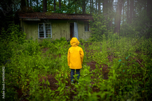 A child in a yellow coat standing in the woods in front of an old wooden house