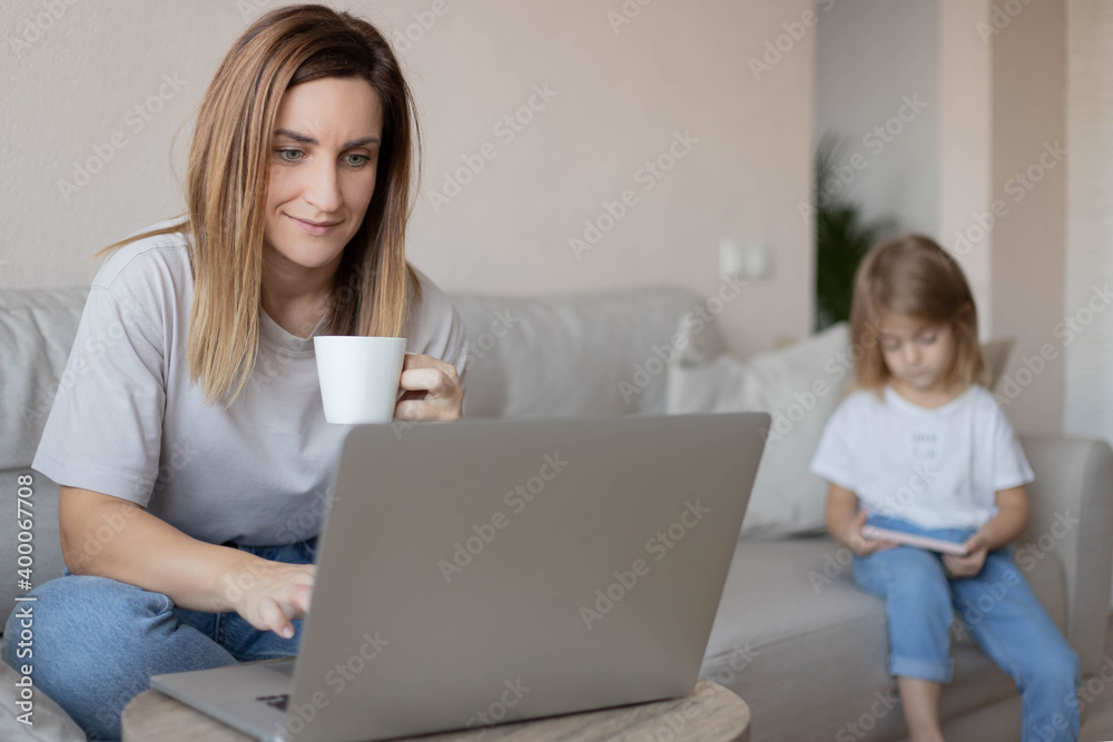 Young beautiful mother businesswoman works at home on the sofa on a laptop, her daughter on the couch sitting with a smartphone. Remote work concept.