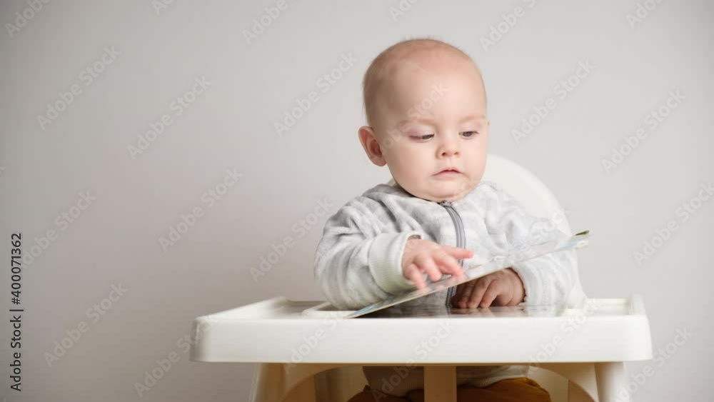Infant boy flips through the pages and looks at the book. Little baby boy sits in baby high chair indoors. Infant boy flips through the pages and looks at the book. Little baby boy sits in baby high chair indoors.