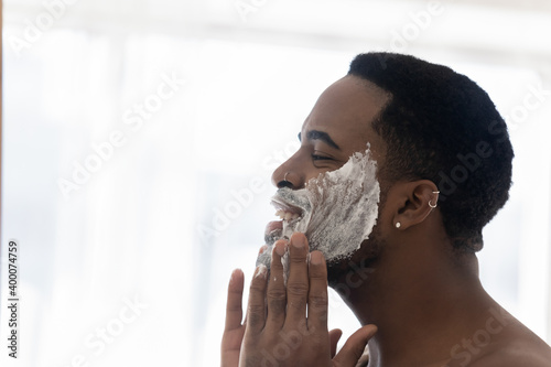 Close up head shot side view happy young african mixed race man cleansing face with foamy scrub, making skincare detox domestic procedures, exfoliating dead cells, enjoying morning hygienic routine.