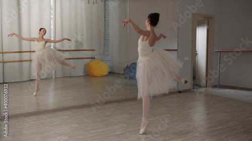 A ballerina in a white ballet suit performs various ballet moves in front of a mirror in the dance hall during a rehearsal photo