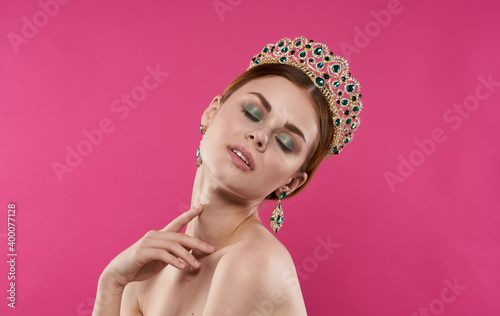 Charming lady with a crown on her head bared shoulders earrings makeup model pink background