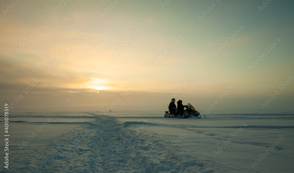 Two riders on snowmobile in sunset light with frozen lake background