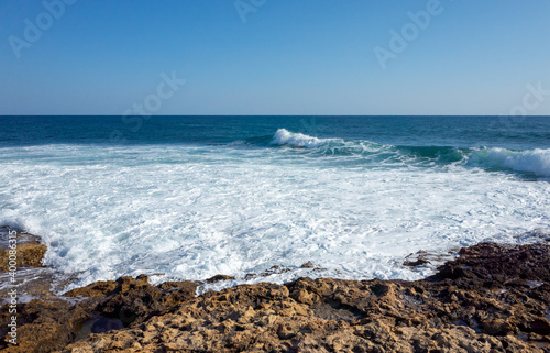 Waves break on the rocky shore, forming mountains of white foam on a clear Sunny day.