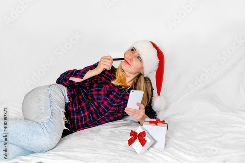 Christmas present. Mischievously smiling woman with gift box, smartphone and redit card wearing santa hat lying on white background. Sales. Christmas and New Year shopping. photo