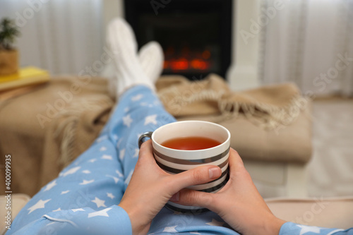 Woman with cup of tea resting near fireplace at home, closeup