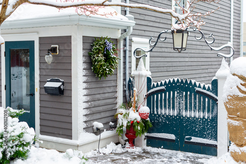 A blue wooden gate with a wrought iron fancy light and decorative gate. There's a Christmas wreath and greenery arrangement near the door of the grey building. Snow covers the ground, fence and shrubs