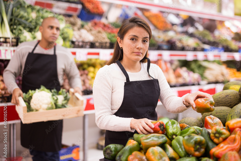 Latin American supermarket woman employee in black apron and male assistant in background