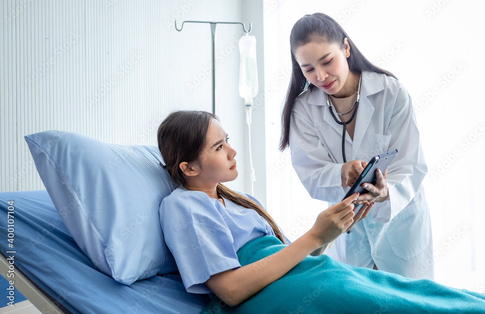 Asian female doctor therapeutic advising with tablet to young female patient on Bed in hospital background.