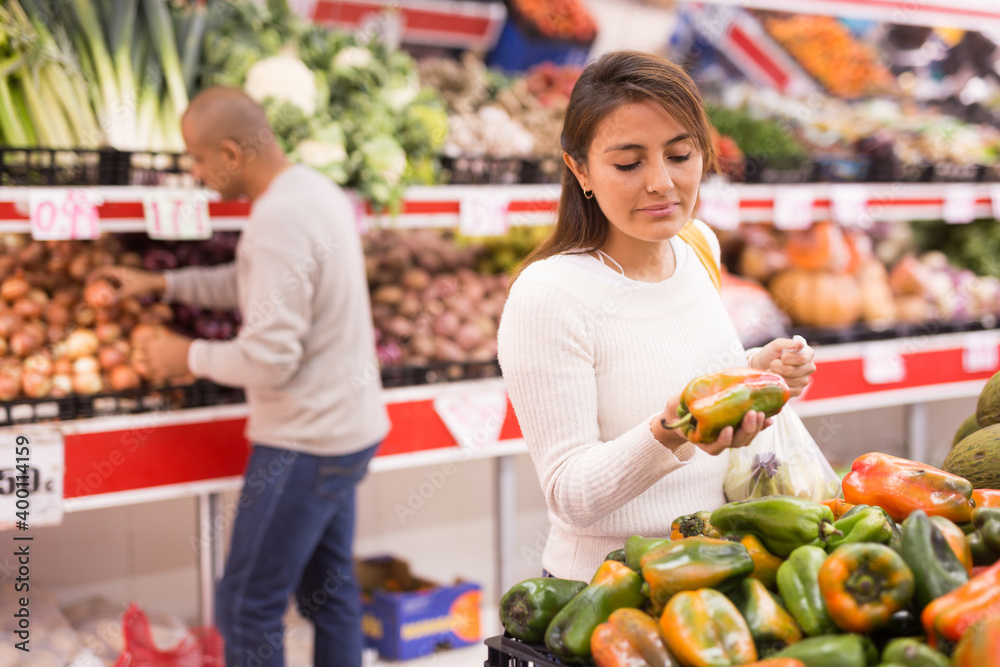 Adult latin american woman choosing sweet pepper in supermarket with other buyer