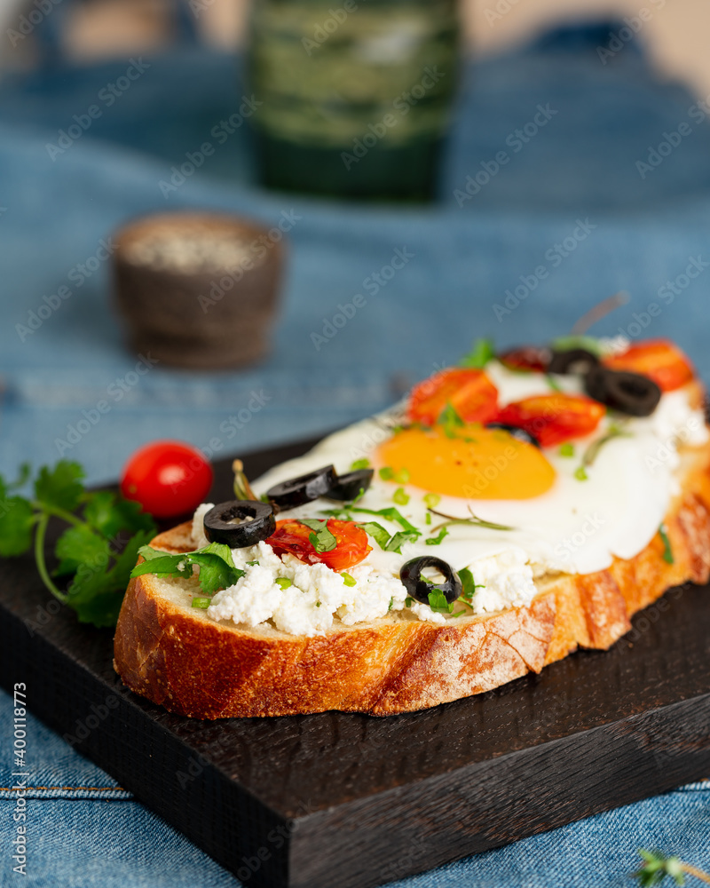 Toasted bread toast with fried eggs with yellow yolk and tomatoes, olives, sprinkled with herbs on dark wooden serving board on blue denim napkin, vertical Toasted bread toast with fried eggs with yellow yolk and tomatoes, olives, sprinkled with herbs on dark wooden serving board on blue denim napkin, vertical