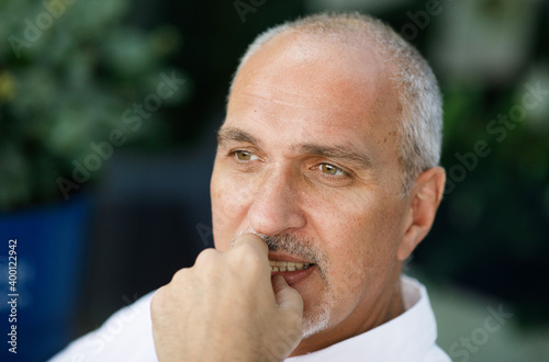  Emotional portrait of a calm and thoughtful mature tanned European man with a bald head looking at tourists sitting in a summer cafe. Summertime. Lifestyle. Summer vacation,. Positive emotions