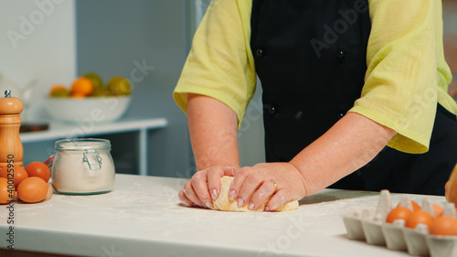Grandmother hands preparing homemade cookies in modern kitchen kneading on the table. Retired elderly baker with bonete mixing ingredients with wheat flour for baking traditional cake and bread