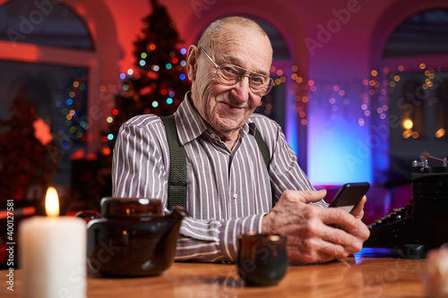 Portrait of a elder cheerful man sitting at table in lovely and colourful room with custom interior decorated with xmas tree and twonkly lights.