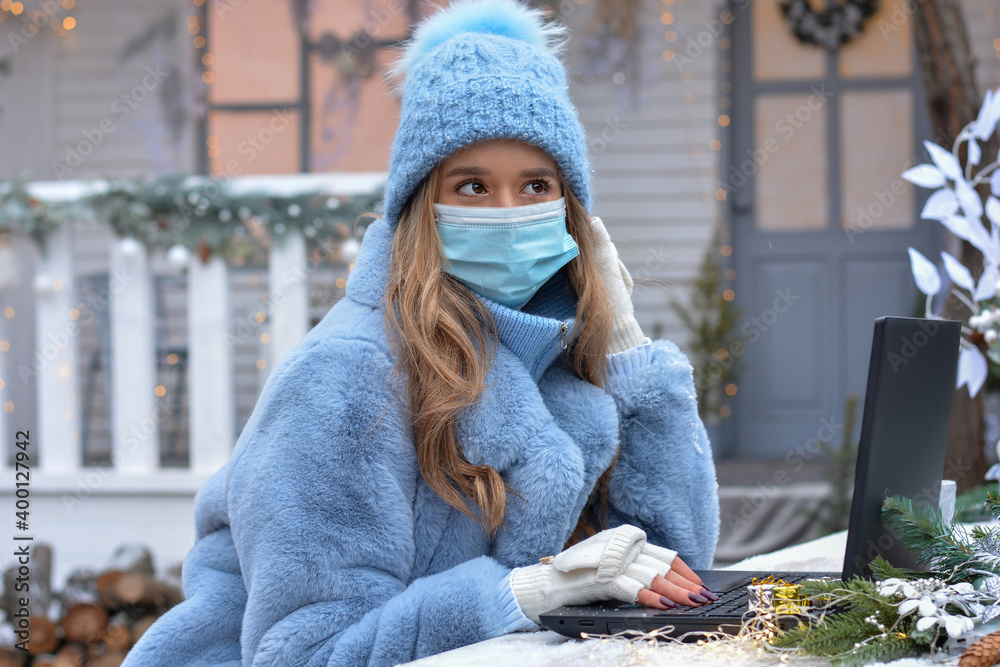 Girl in medical mask communicates with friends on the network through a laptop on the background of a Christmas house. Christmas greetings online,Quarantine, Holidays during the coronavirus outbreak