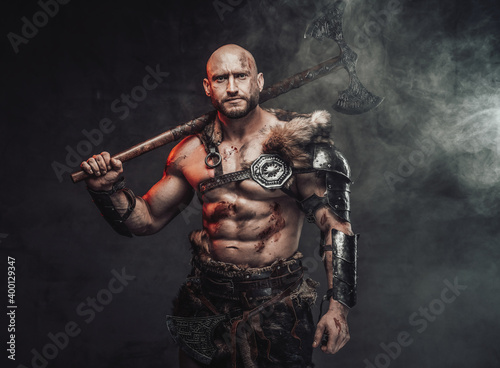 Portrait of a viking warrior with grimy and bloody skin in light armour with fur in dark smokey background holding an axe on his shoulder