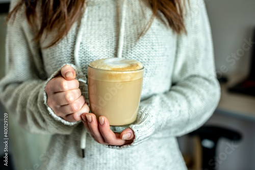 A woman in a warm sweater is holding in her hands a clear glass cup with hot aromatic coffee in her hands. Concept of a cozy morning. Closeup