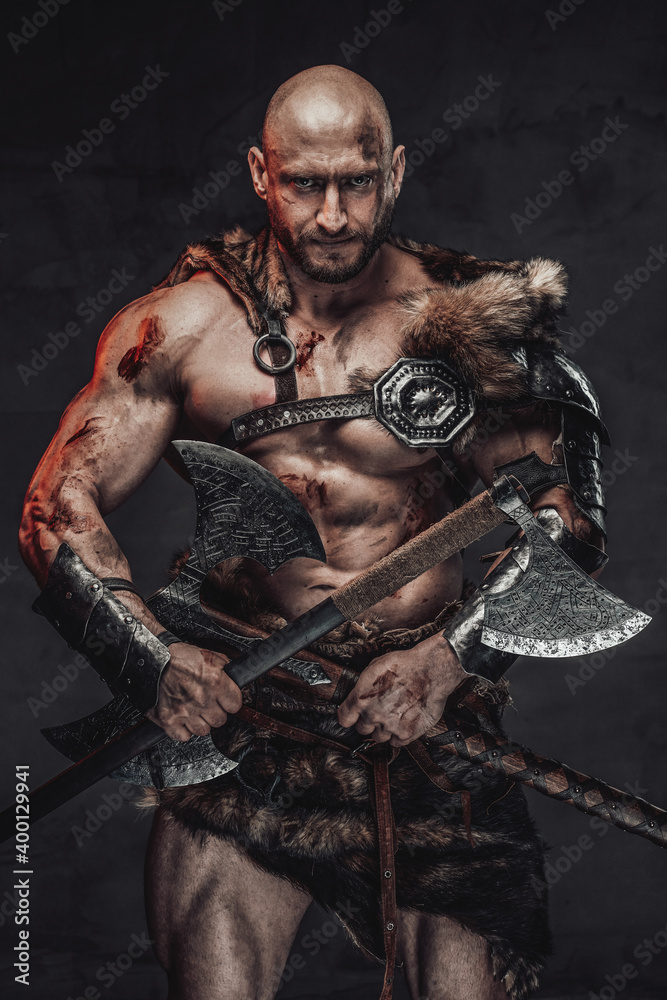 Savage and skilled scandinavian warrior with bald head wielding two axes and in armour with fur in dark background looking at camera with serious face