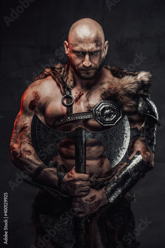 Fototapeta Naklejka Na Ścianę i Meble -  Serious and furious scandinavian barbarian with bald head and grimy and bloody skin in armour with fur in dark background holding two handed double axe