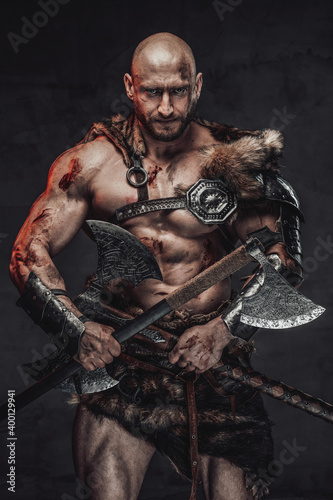 Savage and skilled scandinavian warrior with bald head wielding two axes and in armour with fur in dark background looking at camera with serious face