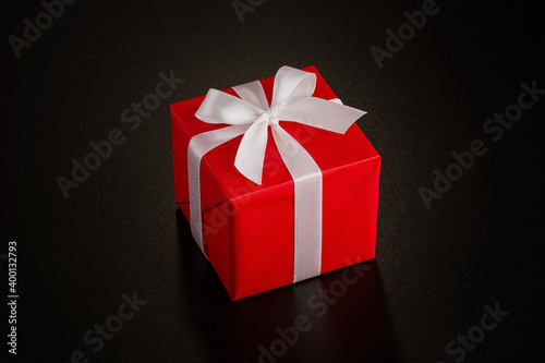 One square gift wrapped in red wrapping paper with white ribbon and bow on a black background with copy space. Close up.