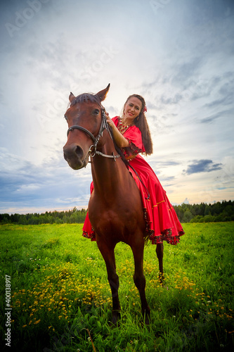 A woman in a bright Gypsy dress and image with a horse in a field with green grass. A model or actress posing in nature with an animal from a farm and the sky with clouds in the background © keleny