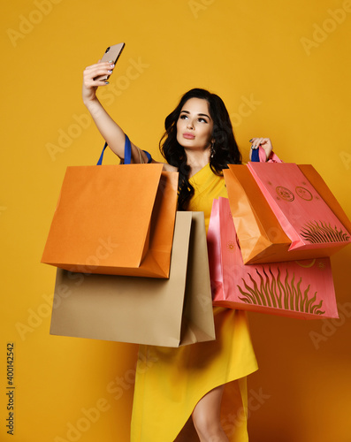 Trendy woman in stylish yellow dress online shopping with modern pink orange and brown shopping bags makes selfie using her mobile smartphone on yellow background. Big Sales