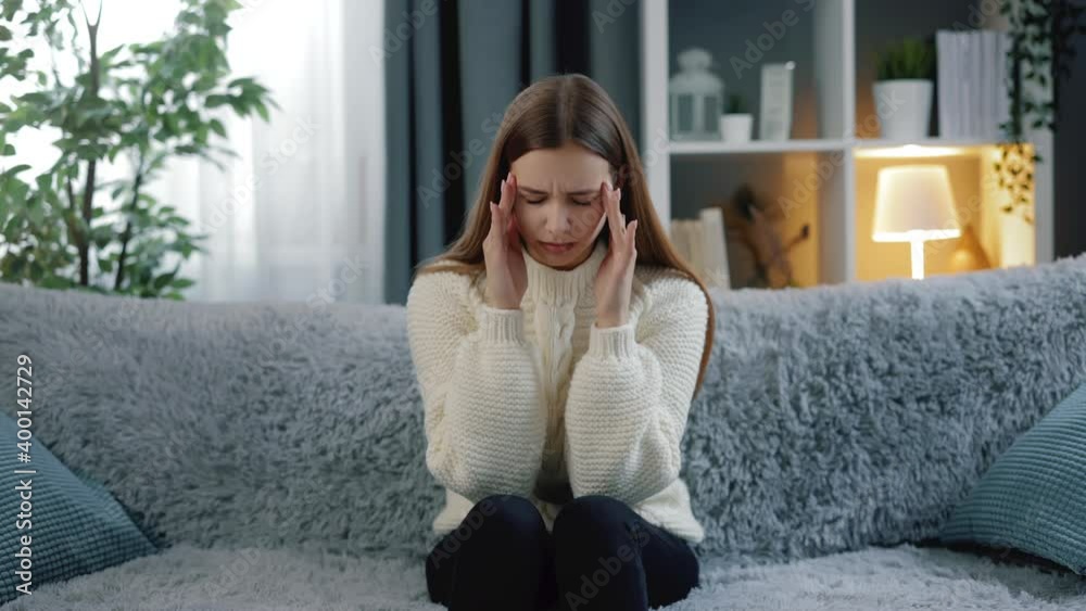 Young beautiful woman in knitted sweater and blue jeans sitting on couch and feeling strong pain in head. Concept of migraine and health care. Young beautiful woman in knitted sweater and blue jeans sitting on couch and feeling strong pain in head. Concept of migraine and health care.