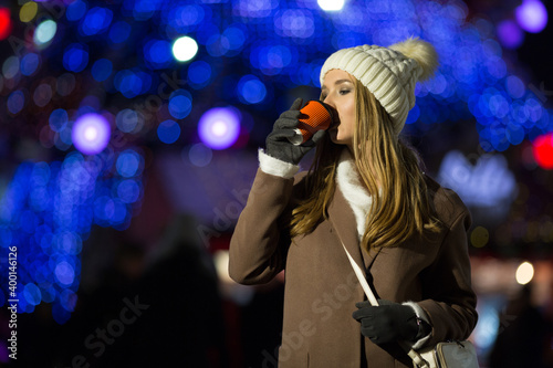 a blonde girl  in the evening  against the background of the lights of the fair with a drink in an orange glass  a white hat and a beige coat walks on the street. smile