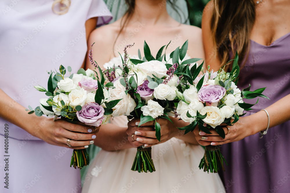 The bride and bridesmaids in an elegant violet dress is standing and holding hand bouquets of pastel pink flowers and greens at nature. Young beautiful girls holds a wedding bouquet outdoors.