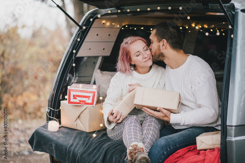Happy young couple in love sitting in a van decorated with festive Christmas lights, hugging and laughing.