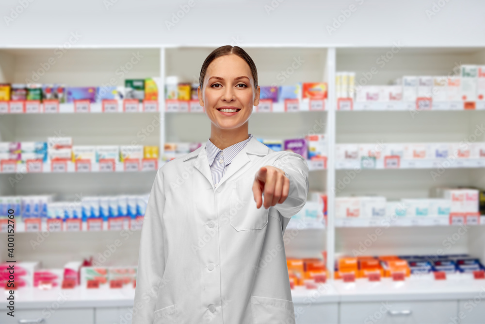 medicine, profession and healthcare concept - happy smiling female pharmacist or doctor in white coat pointing to camera over pharmacy background
