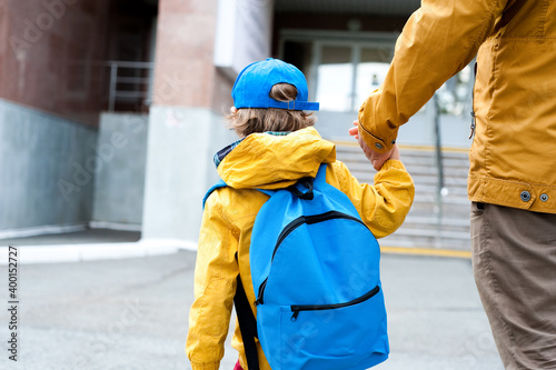 A cute boy in a blue cap and with a blue backpack goes to school with his father.