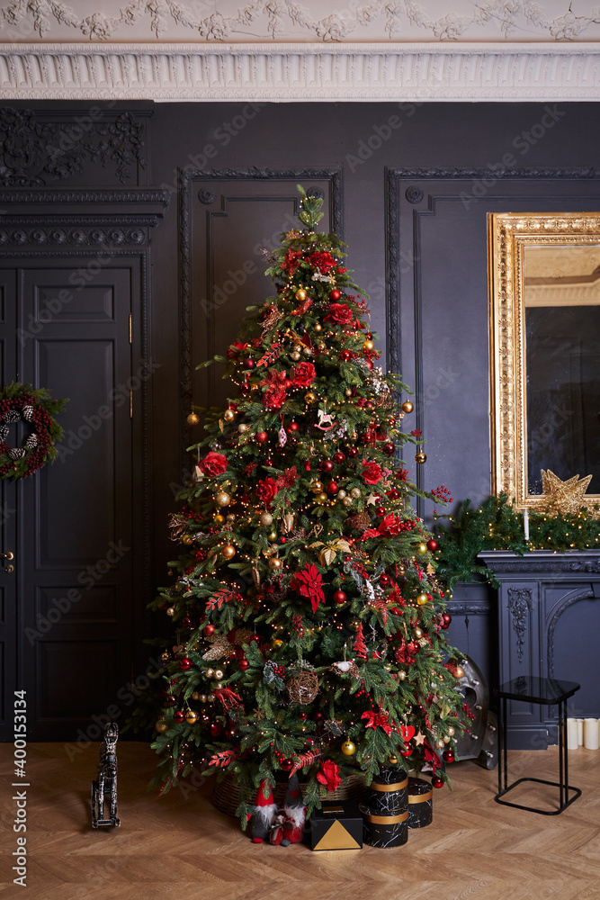 a tall, beautifully decorated Christmas tree stands against the background of a dark gray wall and a mirror in a golden frame, a wreath with snow-covered cones hangs on the doors