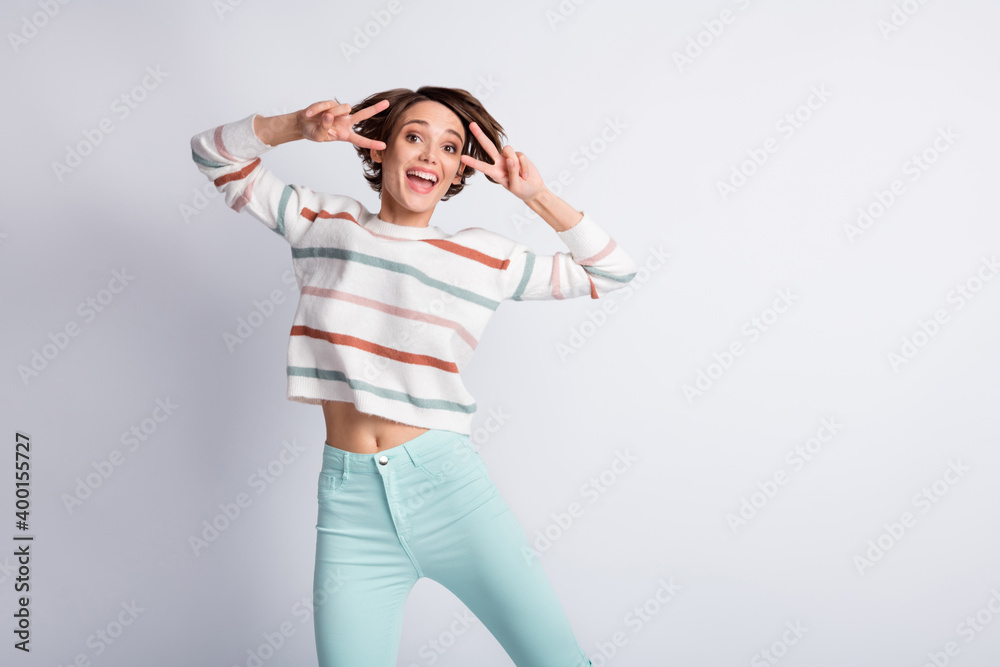 Photo of adorable sweet young lady wear striped sweater dancing showing v-sign isolated grey color background Photo of adorable sweet young lady wear striped sweater dancing showing v-sign isolated grey color background