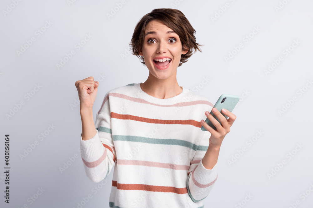 Photo of excited young lady wear striped sweater holding modern gadget rising fist isolated grey color background