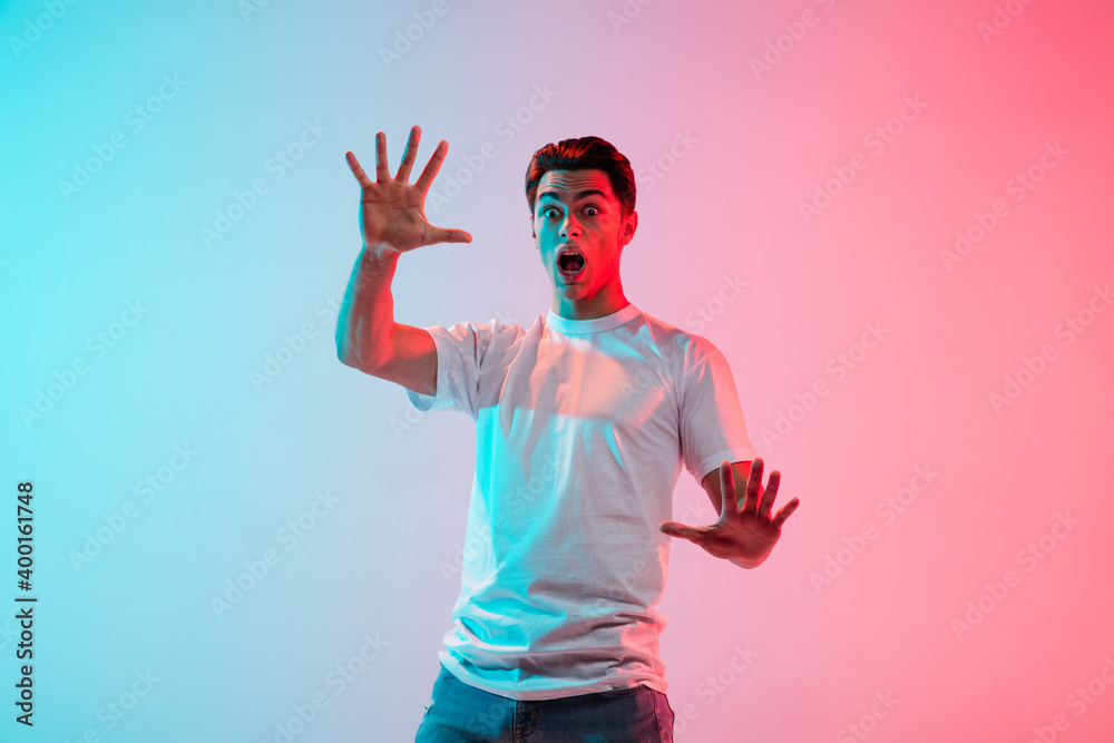 Naklejka premium Scared, shocked. Young caucasian man's portrait on gradient blue-pink studio background in neon light. Concept of youth, human emotions, facial expression, sales, ad. Half length, copyspace.