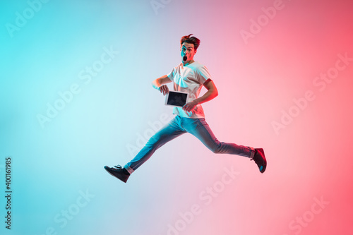 Tablet's screen. Young caucasian man's jumping high on gradient blue-pink studio background in neon light. Concept of youth, human emotions, facial expression, sales, ad. Full length, copyspace.