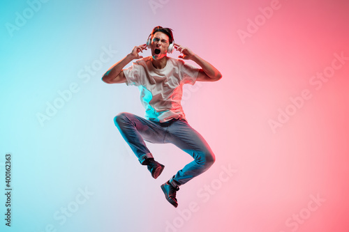 Listening to music. Young caucasian man's jumping on gradient blue-pink studio background in neon light. Concept of youth, human emotions, facial expression, sales, ad. Full length, copyspace.