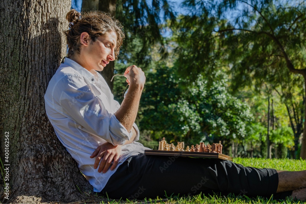 Fototapeta premium Pensive Handsome Chess Player With Long Hair Topknot and White Shirt Playing Chess Alone Sit in the Park