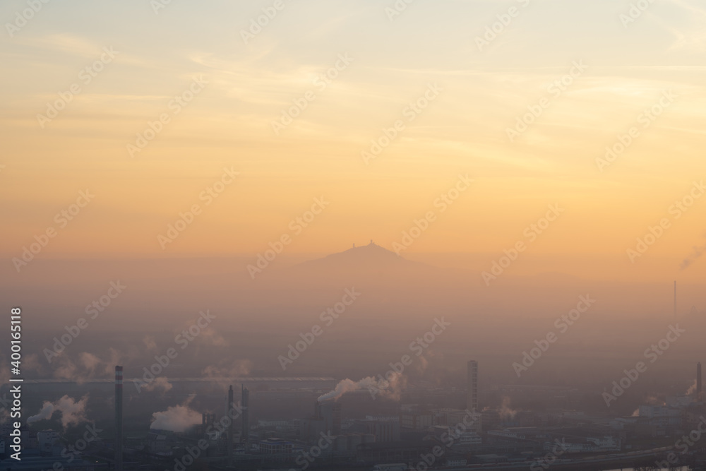 Chemical plant with smoking chimneys in Lovosice, Czech Republic at sunset. Hill with a silhouette of ruins of Hazmburk castle peaking out from low clouds