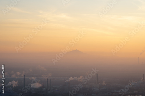Chemical plant with smoking chimneys in Lovosice, Czech Republic at sunset. Hill with a silhouette of ruins of Hazmburk castle peaking out from low clouds