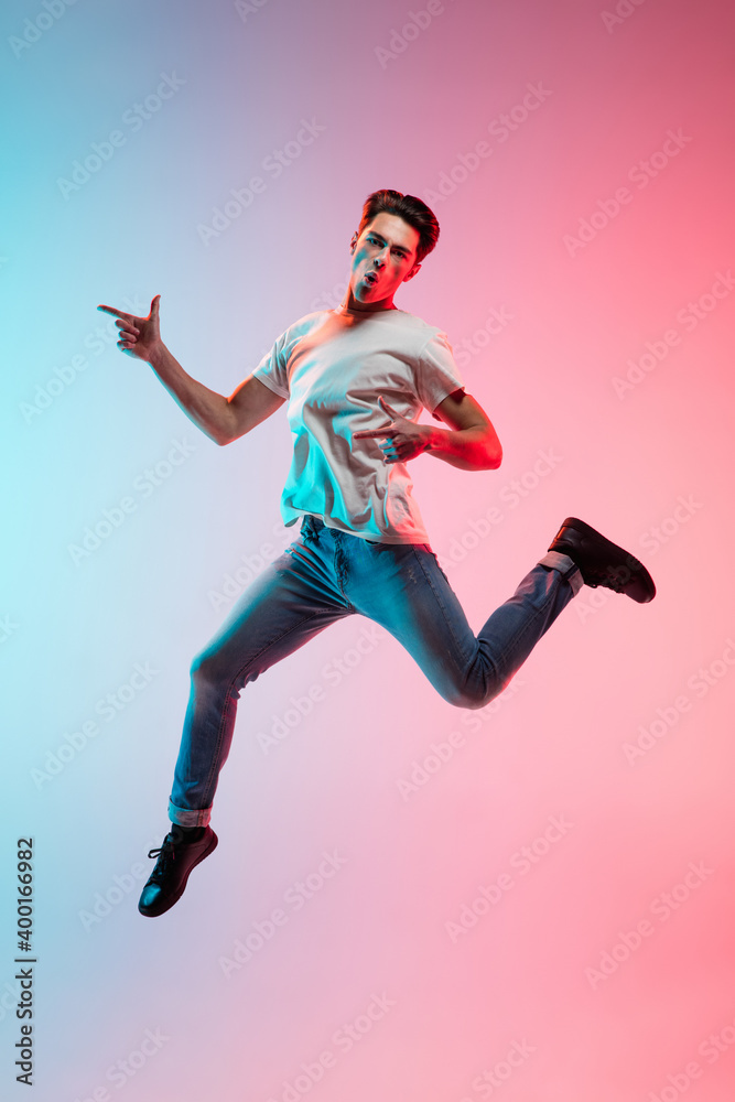 Pointing at side. Young caucasian man's jumping on gradient blue-pink studio background in neon light. Concept of youth, human emotions, facial expression, sales, ad. Full length, copyspace.