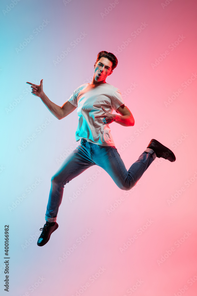 Pointing at side. Young caucasian man's jumping on gradient blue-pink studio background in neon light. Concept of youth, human emotions, facial expression, sales, ad. Full length, copyspace. Pointing at side. Young caucasian man's jumping on gradient blue-pink studio background in neon light. Concept of youth, human emotions, facial expression, sales, ad. Full length, copyspace.