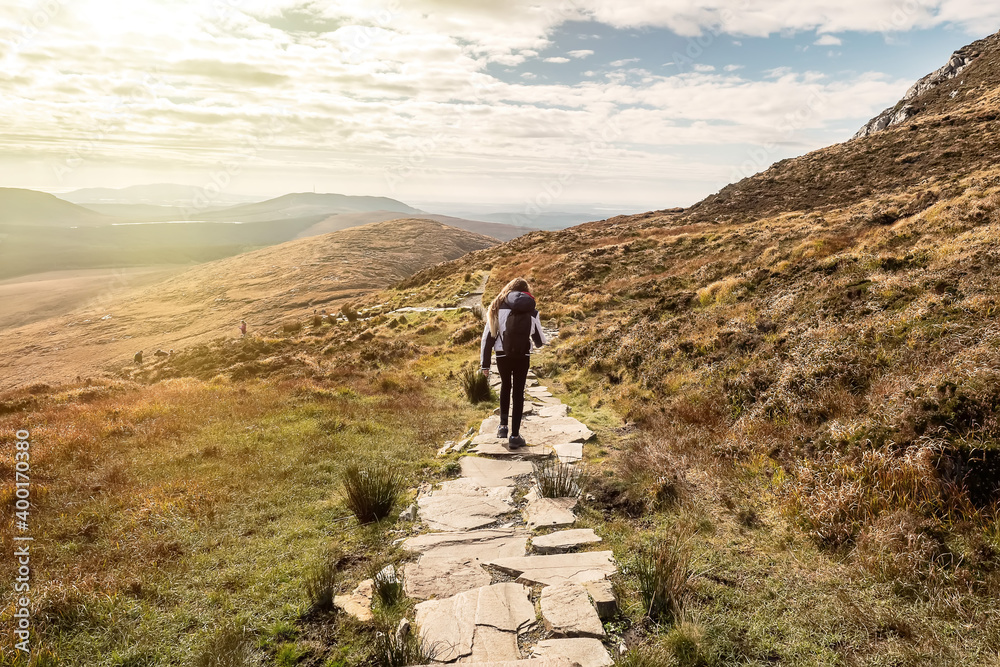 Obraz premium Teenager girl on a hike in a mountains. The model walks on a stone walk path. Letterfrack, Connemara National Park, county Galway, Ireland. Warm sunny day. Travel and hiking concept
