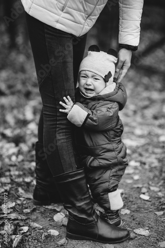 A small child is crying  hugging his mother by the leg. Portrait of a cute boy. Black and white children s photography.
