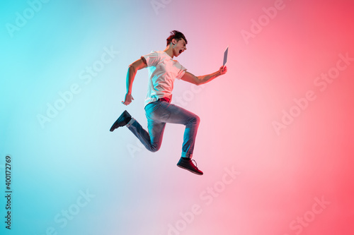 Winner with tablet. Young caucasian man's jumping on gradient blue-pink studio background in neon light. Concept of youth, human emotions, facial expression, sales, ad. Full length, copyspace.
