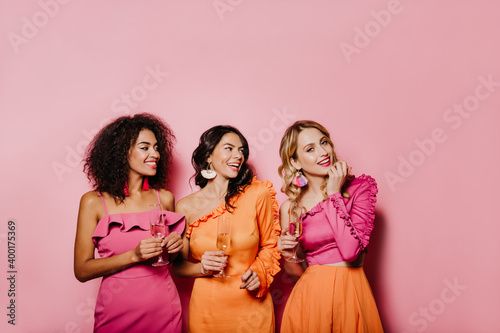 Female models in bright outfits celebrating holidays. Portrait of three women with wineglasses.