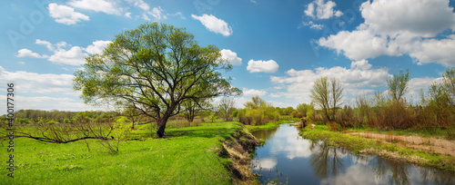 The river among green fields and clouds reflecting in the water in spring at times. fresh spring foliage on the trees. Panoramic photo.