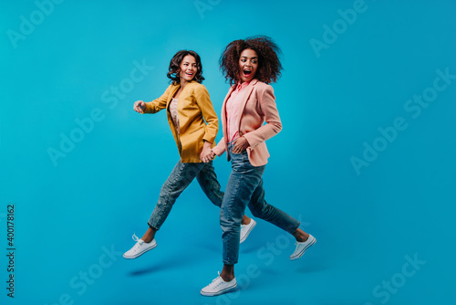 Joyful female models running on blue background. Full-length portrait of two stylish women in jeans.